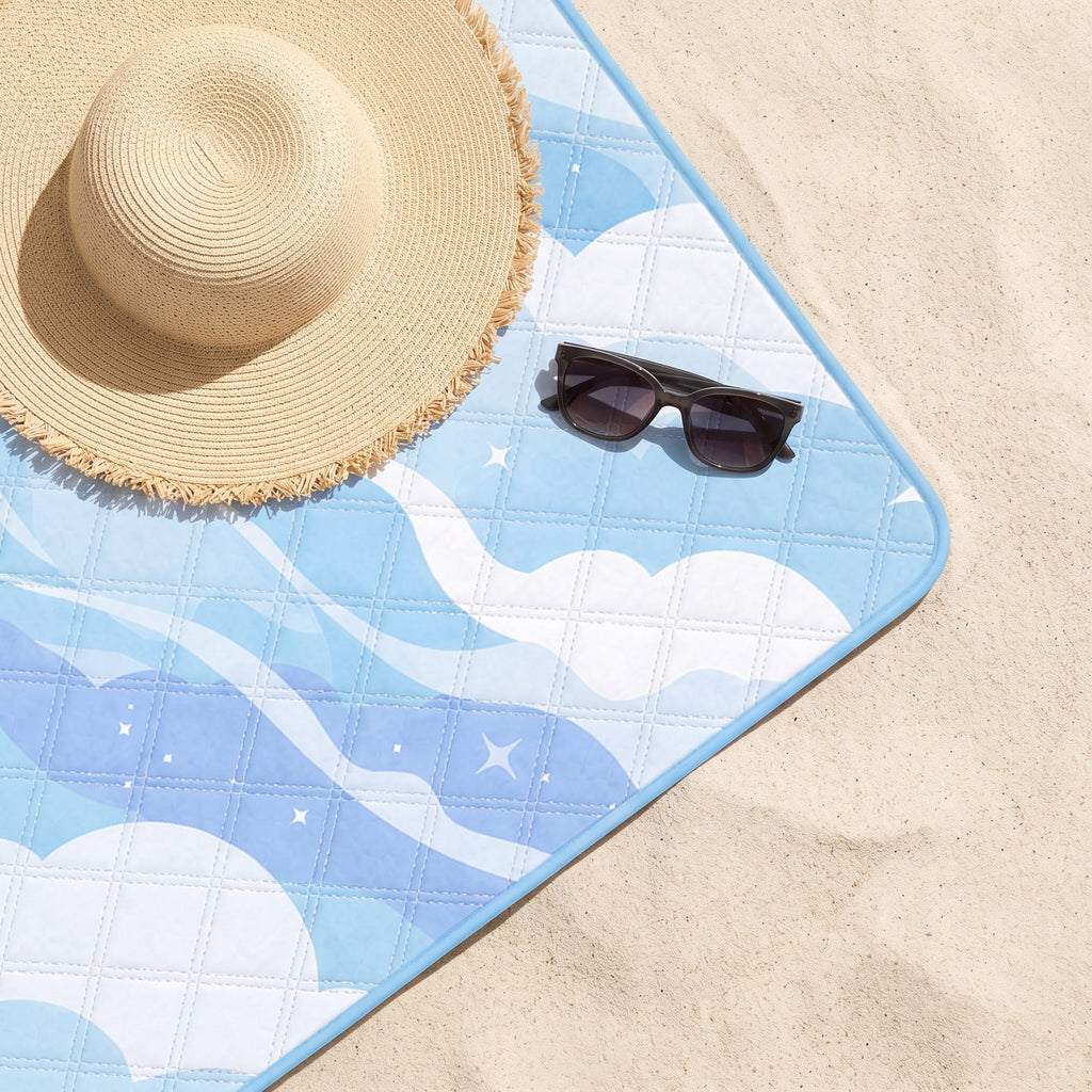 Close-up of the Sky Just Chillin’ quilted beach blanket on sand with a sun hat and sunglasses