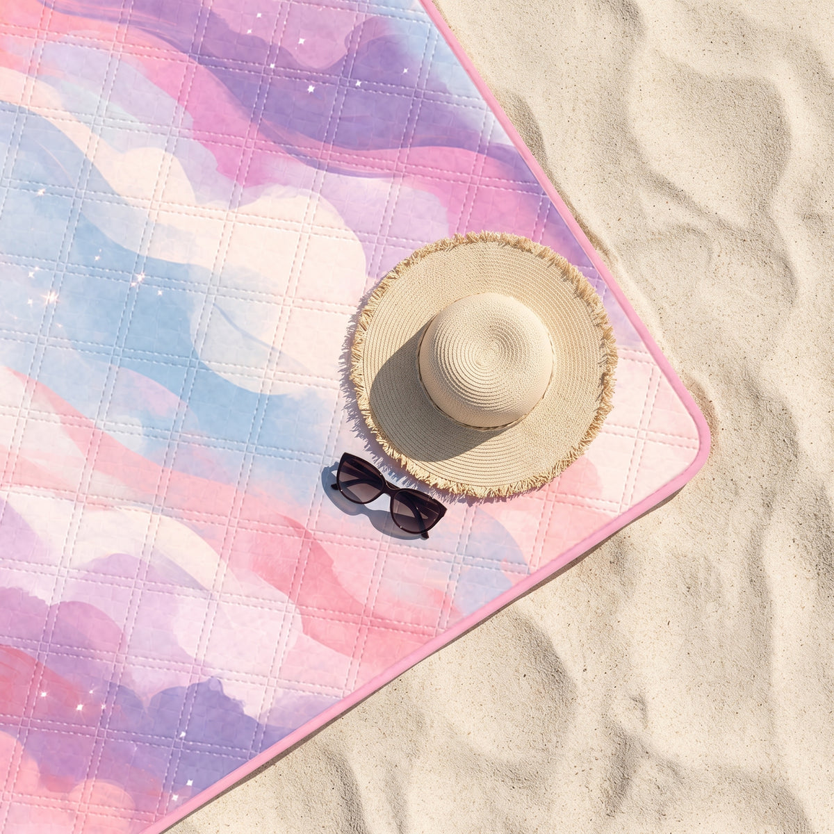 Close-up of the Pink Good Mood Only quilted beach blanket on sand with a sun hat and sunglasses