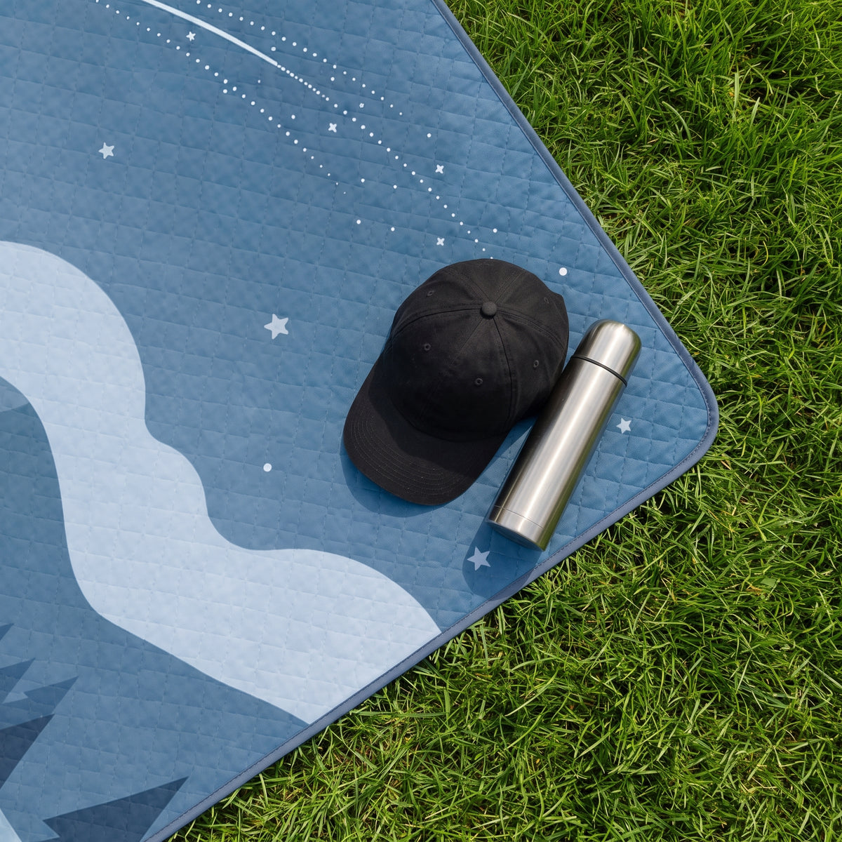 Close-up of the Navy Stay Cozy quilted outdoor blanket on grass with a cap and reusable bottle