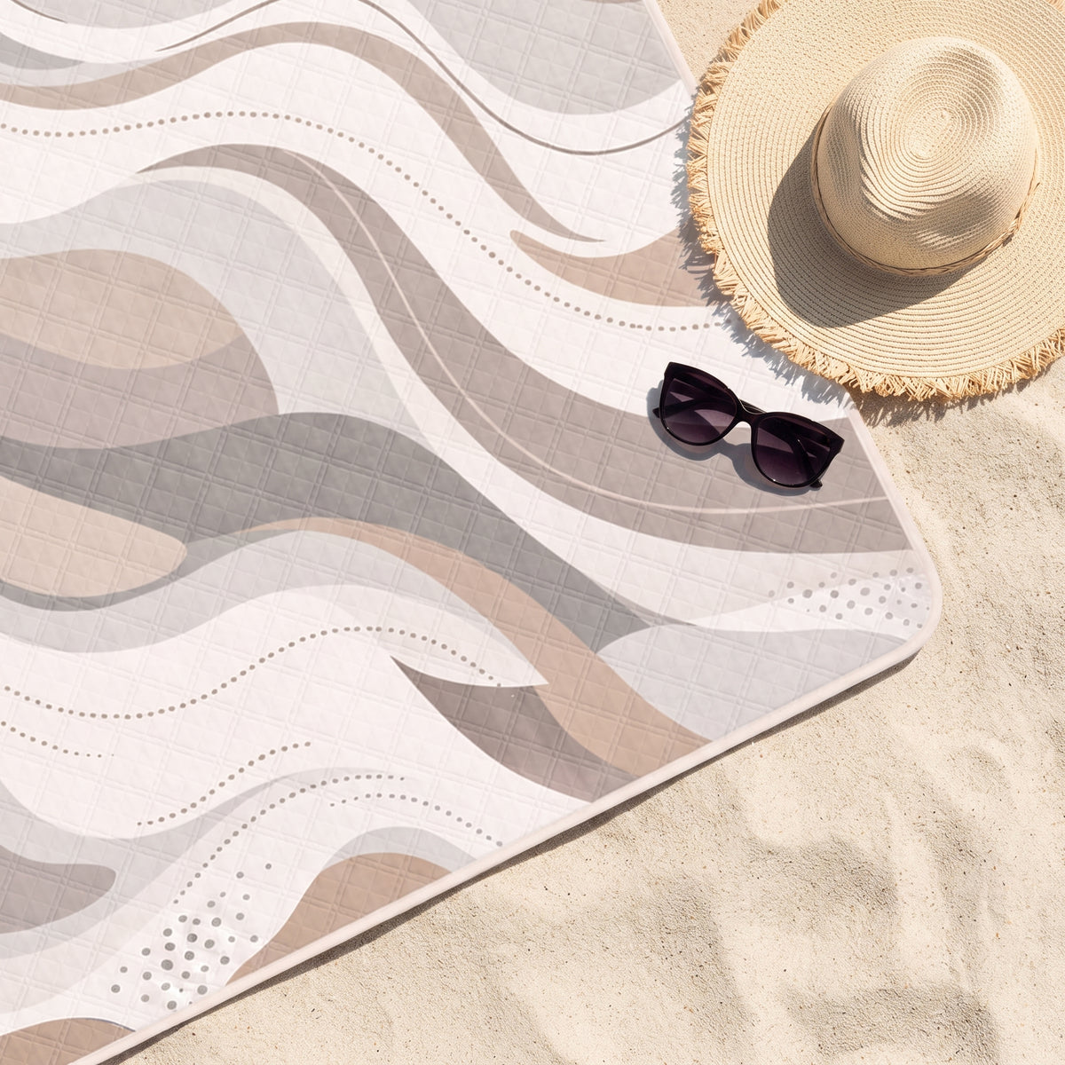 Close-up of the Taupe Take It Easy quilted beach blanket on sand with a sun hat and sunglasses
